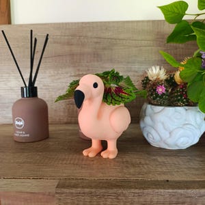 Könnte beinhalten: Ein rosa Flamingo-Pflanztopf mit einer kleinen Pflanze mit grünen und roten Blättern. Der Flamingo hat einen schwarzen Schnabel. Eine braune Diffusorflasche und ein weißer dekorativer Topf mit einem Kaktus sind ebenfalls sichtbar.