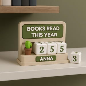 Puede incluir: Un contador de libros beige y verde con el texto "BOOKS READ THIS YEAR" y el nombre "ANNA". El contador muestra el número 255. Un pequeño cactus en una maceta marrón está en el contador, y un cubo separado muestra el número 3.