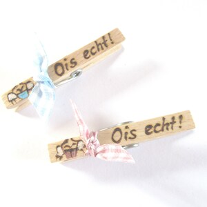 Könnte beinhalten: Zwei Holz-Wäscheklammern mit einer blauen und rosa karierten Schleife. Jede Wäscheklammer hat den Text "Ois echt!" aufgeschrieben.
