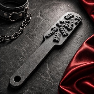 May include: A black paddle featuring a textured surface with several small, raised rectangular brick-like shapes attached to the head. The paddle includes a circular hole at the end of the handle and is accompanied by a black leather collar with a metal chain.