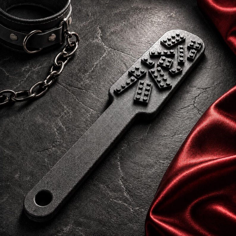 May include: A black paddle featuring a textured surface with several small, raised rectangular brick-like shapes attached to the head. The paddle includes a circular hole at the end of the handle and is accompanied by a black leather collar with a metal chain.