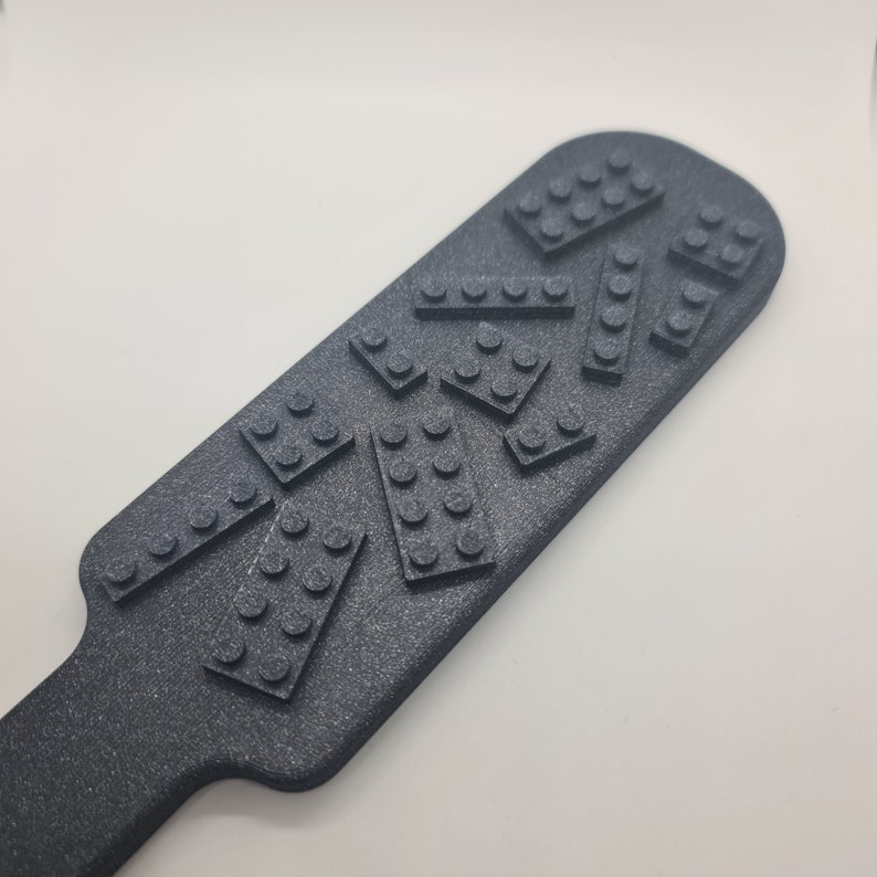 May include: A dark gray, textured paddle featuring a series of raised, rectangular brick-like shapes with circular studs on the surface. The object has a rounded, elongated head and a narrow handle, with a consistent matte finish across the entire piece.