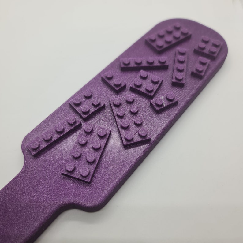 May include: A purple paddle with a textured surface featuring several raised rectangular brick-like shapes with circular studs on top. The object has a rounded end and a narrow handle section, all finished in a consistent matte purple color.