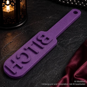 May include: A purple paddle featuring the word BITCH embossed in raised capital letters on the surface. The object has a long handle with a circular hole at the end for hanging. The texture of the paddle appears matte and uniform in colour.