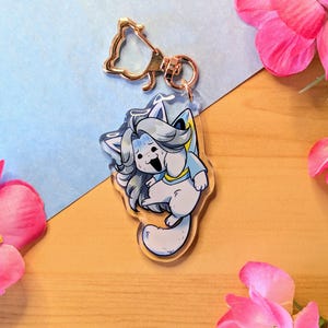 Puede incluir: Un llavero acrílico transparente con un personaje de gato de dibujos animados con pelaje gris, ropa azul y una expresión angustiada. El llavero tiene un cierre dorado y está sobre un fondo azul y de madera.