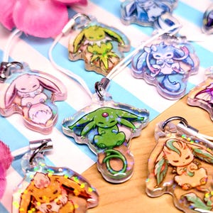 May include: A collection of clear acrylic charms featuring various colorful cartoon creatures with holographic glitter accents. Each charm is attached to a white cord loop and displays a unique character design, including pink, green, blue, and orange figures.