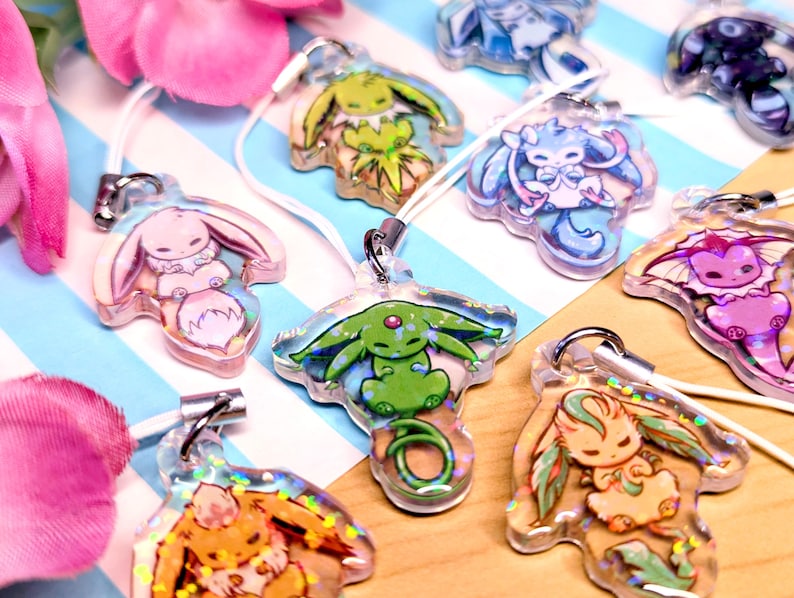 May include: A collection of clear acrylic charms featuring various colorful cartoon creatures with holographic glitter accents. Each charm is attached to a white cord loop and displays a unique character design, including pink, green, blue, and orange figures.
