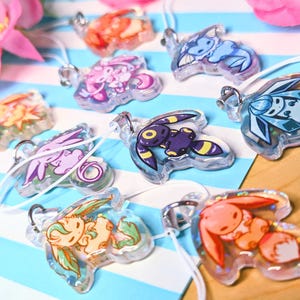 May include: A collection of clear acrylic charms featuring colorful cartoon creature illustrations. Each charm has a silver metal jump ring and a white cord loop. The designs include various characters in shades of blue, purple, orange, and yellow.