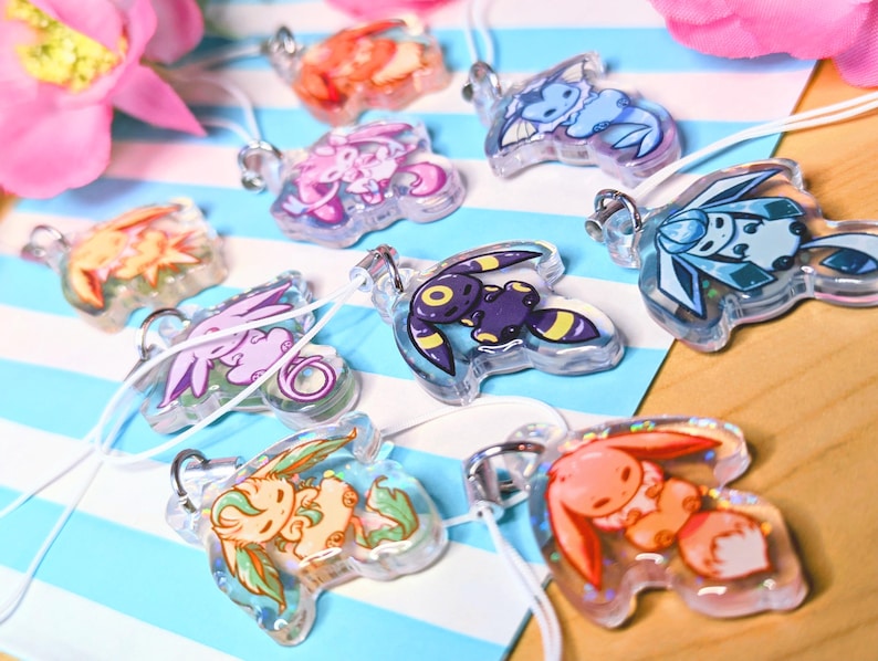 May include: A collection of clear acrylic charms featuring colorful cartoon creature illustrations. Each charm has a silver metal jump ring and a white cord loop. The designs include various characters in shades of blue, purple, orange, and yellow.