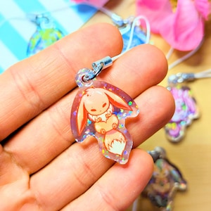 May include: A clear acrylic charm featuring a cartoon creature with long ears and a fluffy tail, embedded with iridescent glitter. The charm is shaped like a leaf and includes a silver metal jump ring attached to a white cord for hanging.