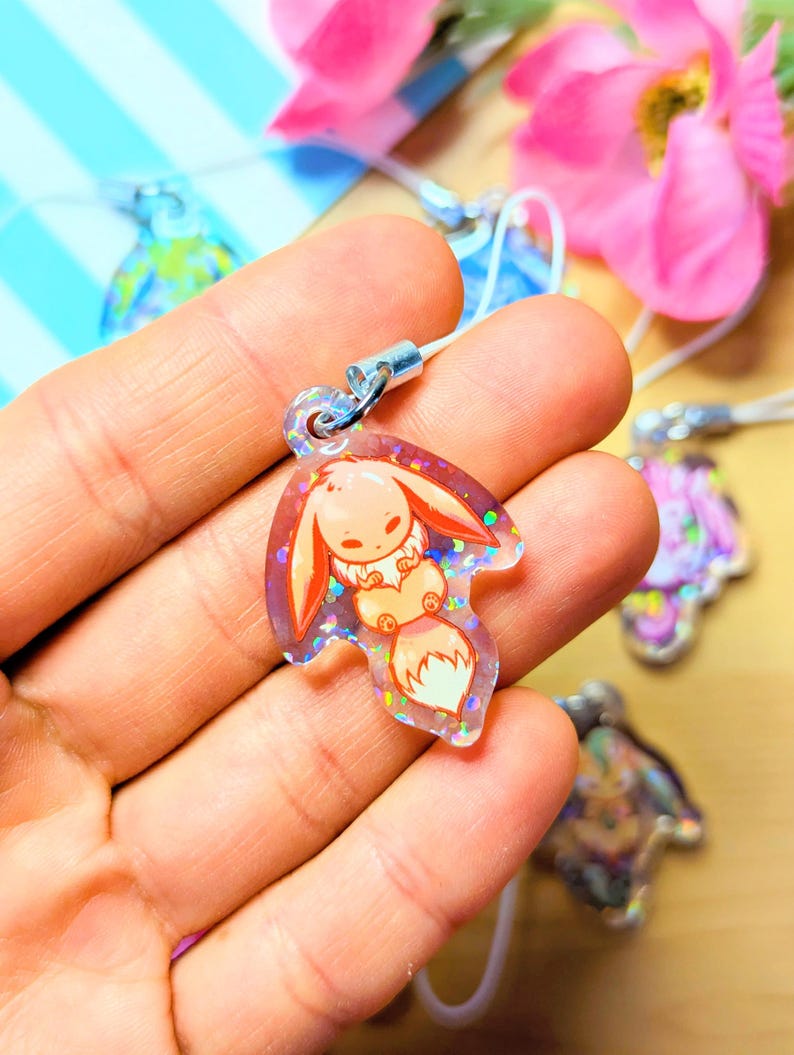 May include: A clear acrylic charm featuring a cartoon creature with long ears and a fluffy tail, embedded with iridescent glitter. The charm is shaped like a leaf and includes a silver metal jump ring attached to a white cord for hanging.