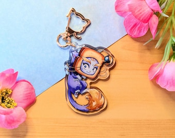 May include: A blue and purple cat with a golden tail is holding a slice of toast with a blue eye on top. The cat is wearing a gold headband and is attached to a gold keychain.
