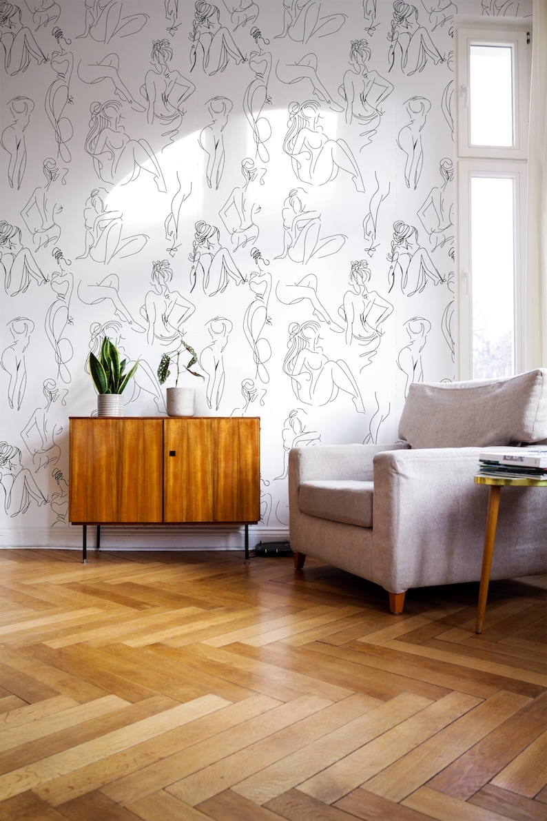 May include: A white wall with a black line drawing pattern of women's figures. The wall is in a living room with a wooden cabinet, a grey armchair, and a wooden end table with a stack of books.