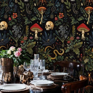 Op de afbeelding: Een donkere eetkamer met een zwarte behang met een herhalend patroon van schedels, paddenstoelen, slangen en bloemen. Een houten tafel is gedekt met witte borden, gouden onderborden en heldere glazen. Twee houten stoelen zijn zichtbaar.