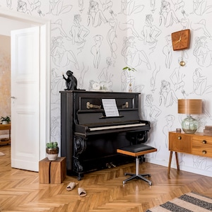 May include: A black upright piano with a wooden stool in a room with white walls featuring a wallpaper pattern of black line drawings of women. A wooden side table with a lamp and a potted plant are also in the room.