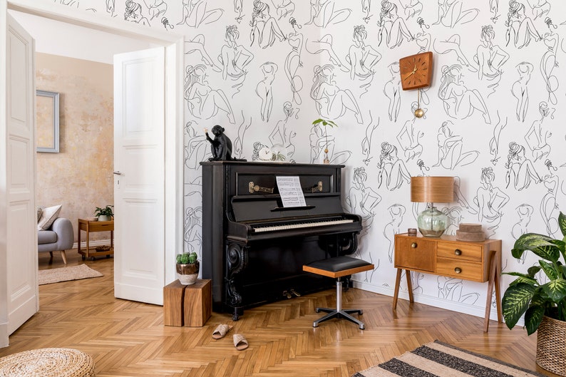 May include: A black upright piano with a wooden stool in a room with white walls featuring a wallpaper pattern of black line drawings of women. A wooden side table with a lamp and a potted plant are also in the room.