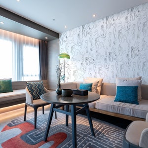 May include: A living room interior with a light gray couch, two armchairs, a coffee table, and a rug. The wall behind the couch is covered in a white wallpaper with a pattern of black line drawings of people. The room has large windows with white curtains.