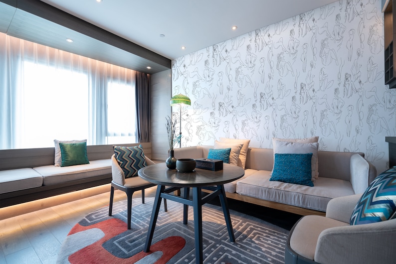 May include: A living room interior with a light gray couch, two armchairs, a coffee table, and a rug. The wall behind the couch is covered in a white wallpaper with a pattern of black line drawings of people. The room has large windows with white curtains.