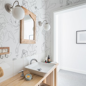 May include: A white bathroom with a white sink and a wooden countertop. The walls are decorated with a black and white line drawing wallpaper of women. There is a mirror above the sink and a small picture frame on the wall.