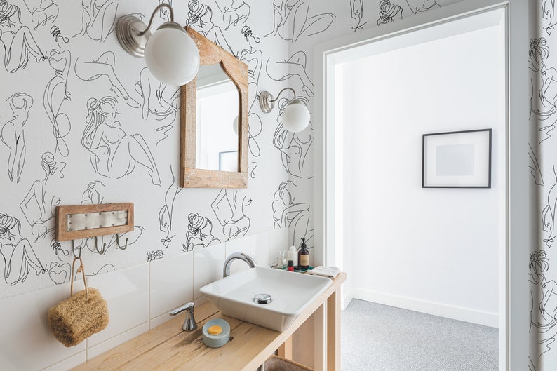 May include: A white bathroom with a white sink and a wooden countertop. The walls are decorated with a black and white line drawing wallpaper of women. There is a mirror above the sink and a small picture frame on the wall.
