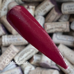 May include: A deep red nail polish with a glossy finish. The polish is shown against a background of wine corks.
