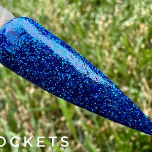 May include: A close-up of a dark blue nail polish with a glittery finish. The polish is applied to a nail tip and the background is blurred green grass.