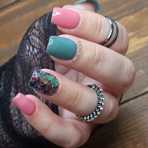 May include: A hand with manicured nails in various colors and designs. The nails feature a matte teal, glitter, and pink polish. The index finger has a glitter design with green, purple, and gold flakes. The ring finger has a matte teal polish. The middle and pinky fingers are painted pink.