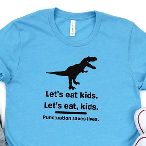 Puede incluir: Una camiseta azul claro con una silueta negra de un dinosaurio y el texto "Let's eat kids. Let's eat, kids. Punctuation saves lives."
