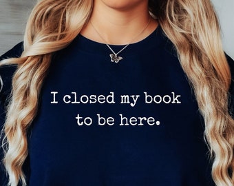 May include: A navy blue crewneck sweatshirt featuring the white text "I closed my book to be here." The garment has long sleeves with ribbed cuffs and a ribbed neckline, constructed from a solid-colored fabric with a simple, clean design.