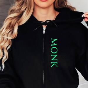 Puede incluir: Sudadera con capucha negra con cierre de cremallera y la palabra "MONK" impresa verticalmente en verde en la parte delantera.
