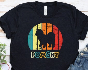 May include: A dark blue t-shirt with a vintage-style graphic of a Pomsky dog in front of a sunset. The text "POMSKY" is printed below the graphic.