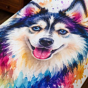 Pode incluir: Uma manta de cama branca com uma grande ilustração colorida em estilo aguarela de um cão husky com olhos azul-brilhantes, cara preta e branca e língua cor-de-rosa, rodeado por salpicos vibrantes de tinta roxa, azul, amarela e rosa.