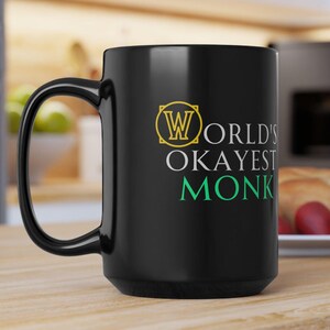 Puede incluir: Taza de café de cerámica negra con un diseño blanco que dice "World's Okayest Monk" en una fuente dorada y verde.