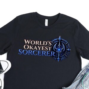 以下が含まれることがあります： 「WORLD'S OKAYEST SORCERER」という白い文字と青い文字がプリントされた黒い半袖Tシャツ。青い円形の魔法のエンブレムが添えられています。Tシャツの横には、黒いキャンバス地のスニーカーと青いデニムのショートパンツが置かれています。