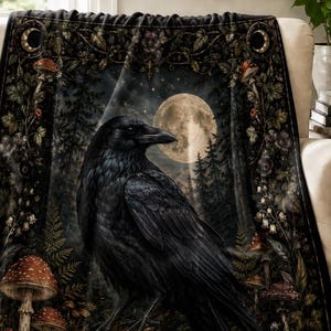 May include: A black throw blanket featuring a detailed illustration of a raven perched on a branch in a dark forest. The design includes a large full moon, numerous red-capped mushrooms, ferns, and a skull at the bottom, framed by a decorative border.