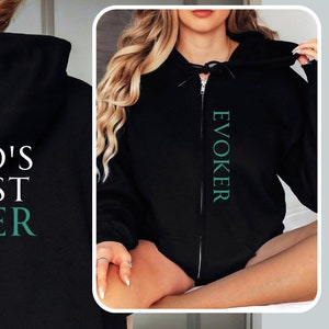 Könnte beinhalten: Schwarzer Kapuzenpullover mit dem Text "World's Okayest Evoker" in gelber und grüner Schrift. Das Wort "Evoker" ist auch vertikal entlang des Reißverschlusses eines schwarzen Kapuzenpullovers gedruckt.
