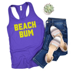 May include: Purple tank top with the text "BEACH BUM" in yellow outlined letters. The tank top is paired with blue jeans and white and pink striped flats.