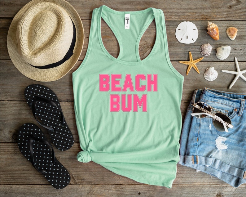 May include: A light green tank top with the words "Beach Bum" in pink letters. The tank top is laying on a wooden surface with a straw hat, flip flops, denim shorts, sunglasses, and seashells.