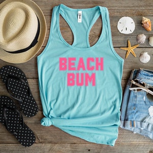 May include: A light blue tank top with the text "BEACH BUM" in hot pink. The tank top is laid out on a wooden surface with a straw hat, flip flops, denim shorts, sunglasses, and seashells.