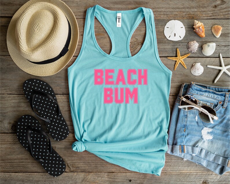 May include: A light blue tank top with the text "BEACH BUM" in hot pink. The tank top is laid out on a wooden surface with a straw hat, flip flops, denim shorts, sunglasses, and seashells.