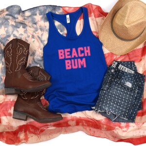 May include: A blue tank top with the words "Beach Bum" in pink letters on a white American flag background. The tank top is paired with denim shorts, cowboy boots, and a straw hat.