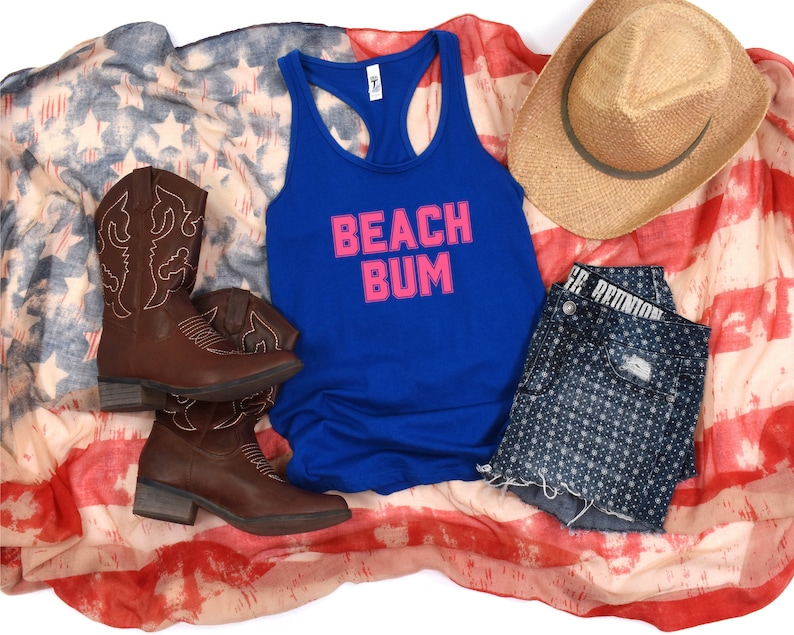 May include: A blue tank top with the words "Beach Bum" in pink letters on a white American flag background. The tank top is paired with denim shorts, cowboy boots, and a straw hat.