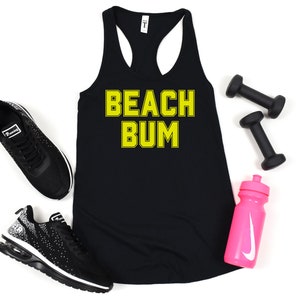 May include: Black tank top with the text "BEACH BUM" in yellow letters. The tank top is laying flat on a white surface with a pair of black sneakers, two black dumbbells, and a pink water bottle with a white Nike swoosh.