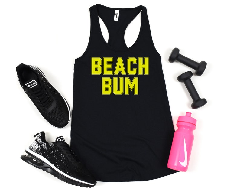 May include: Black tank top with the text "BEACH BUM" in yellow letters. The tank top is laying flat on a white surface with a pair of black sneakers, two black dumbbells, and a pink water bottle with a white Nike swoosh.