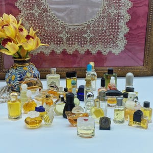 K&ouml;nnte beinhalten: Eine Sammlung von Miniatur-Parf&uuml;mflaschen in verschiedenen Formen und Gr&ouml;&szlig;en, mit Farben von klar bis bernsteinfarben und schwarz. Eine Vase mit gelben Orchideen und eine Perlenkette sind ebenfalls vorhanden.