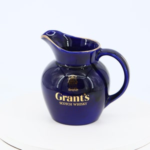 Puede incluir: Una jarra de cerámica azul marino con asa y pico vertedor. La jarra tiene ribetes dorados y las palabras "Grant's SCOTCH WHISKY" en letras doradas. La jarra está sobre una superficie blanca.