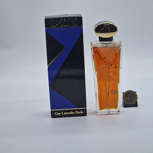 Può includere: Un set di bottiglia e scatola di profumo. La bottiglia di profumo è trasparente con un tappo dorato e contiene un liquido ambrato chiaro. La scatola è nera con un design geometrico blu e oro e il testo "CLANDESTINE" e "Guy Laroche Paris".