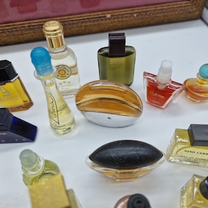 K&ouml;nnte beinhalten: Eine Sammlung von Miniatur-Parf&uuml;mflaschen in verschiedenen Formen und Farben. Die Flaschen bestehen aus klarem, goldenem und farbigem Glas mit unterschiedlichen Verschlussdesigns. Einige Flaschen haben Etiketten mit Text. Die Anordnung befindet sich auf einer wei&szlig;en Oberfl&auml;che.