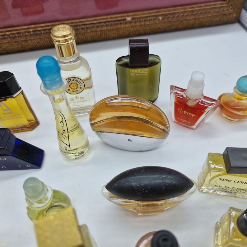 K&ouml;nnte beinhalten: Eine Sammlung von Miniatur-Parf&uuml;mflaschen in verschiedenen Formen und Farben. Die Flaschen bestehen aus klarem, goldenem und farbigem Glas mit unterschiedlichen Verschlussdesigns. Einige Flaschen haben Etiketten mit Text. Die Anordnung befindet sich auf einer wei&szlig;en Oberfl&auml;che.