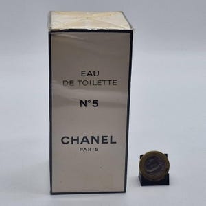 May include: A rectangular perfume box with the text "EAU DE TOILETTE N&deg;5 CHANEL PARIS" in black. The box is beige with a clear plastic wrap. A small coin is next to the box.